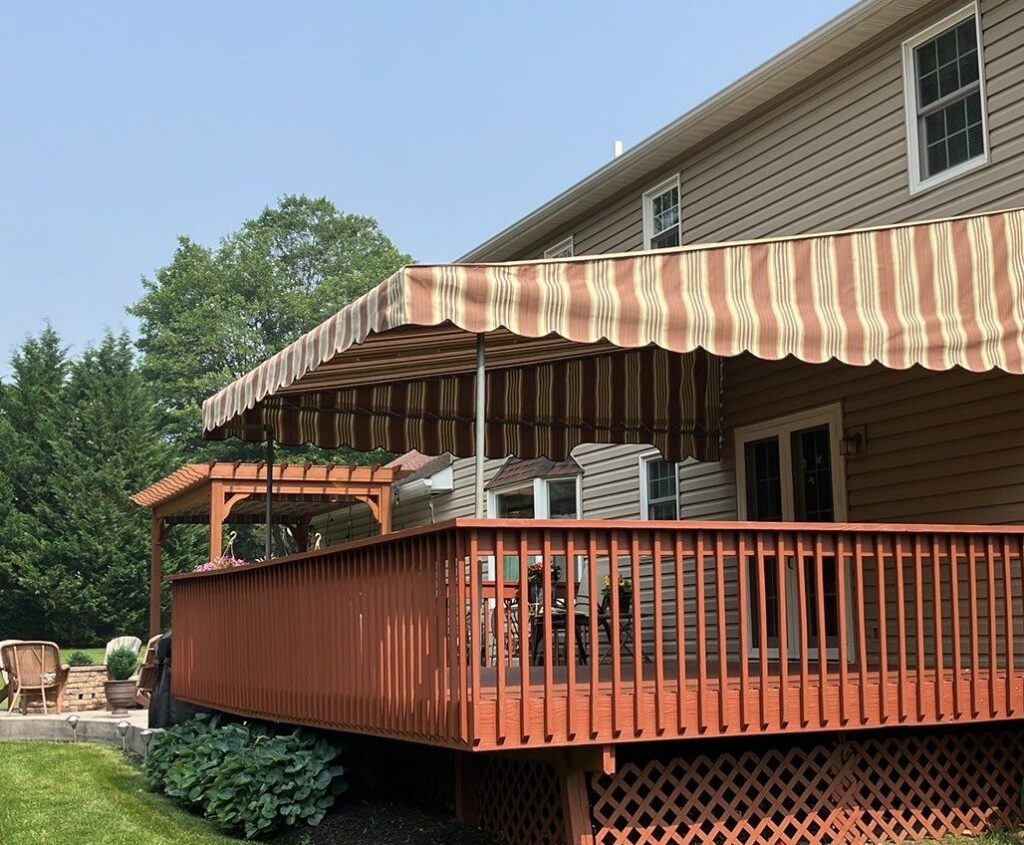 A white and orange striped stationary awning covering a red wooden deck on a clear summer day.