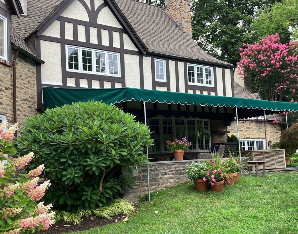 Green stationary awning attached to side of a home, surrounded by a well maintained garden.
